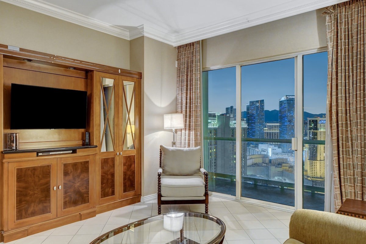 The living area is furnished with a comfortable armchair and a round glass table, bathed in natural light from the large sliding glass doors. The city skyline is visible beyond the balcony, complemented by soft furnishings and elegant decor.