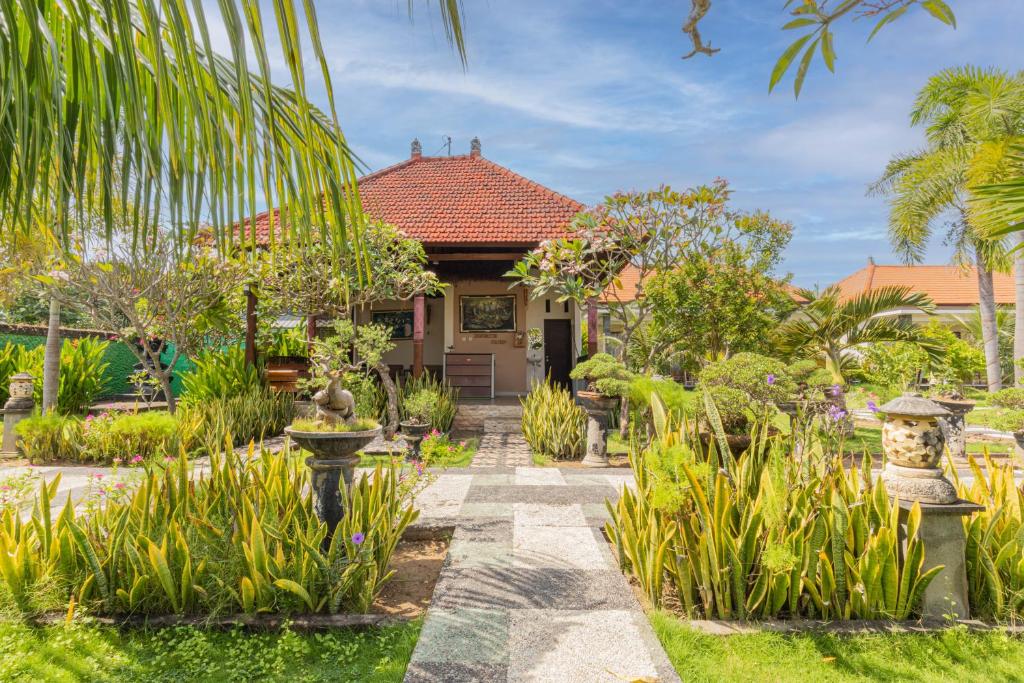 The entrance to the bungalow is surrounded by lush greenery and decorative plants. A stone pathway leads to a traditional roof structure, providing a welcoming view. The serene garden features various flora under a bright sky, accentuating the tranquility of the tropical setting.