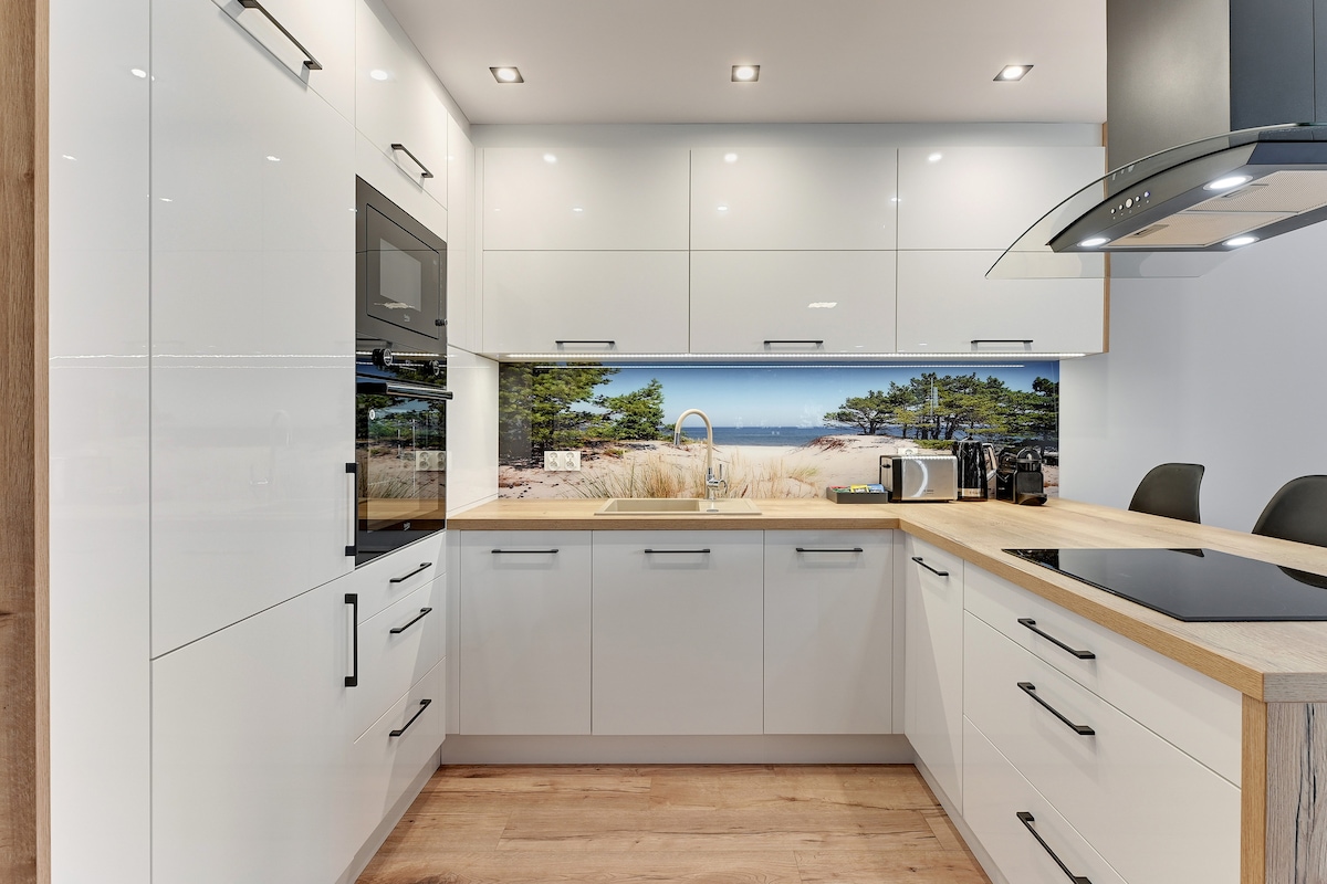 A modern kitchen is displayed, characterized by sleek white cabinetry and a light wood countertop. Appliances include an oven, microwave, and kettle, creating a practical cooking space. A scenic backsplash image features a beach view, enhancing the room's aesthetic with natural elements.