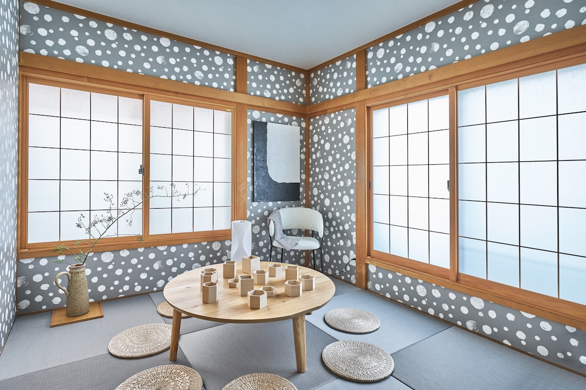 A relaxing room features light grey walls adorned with white dotted patterns. Large windows provide natural light, and a round wooden table is surrounded by several seat cushions. A simple vase with a branch adds a touch of nature to the space.