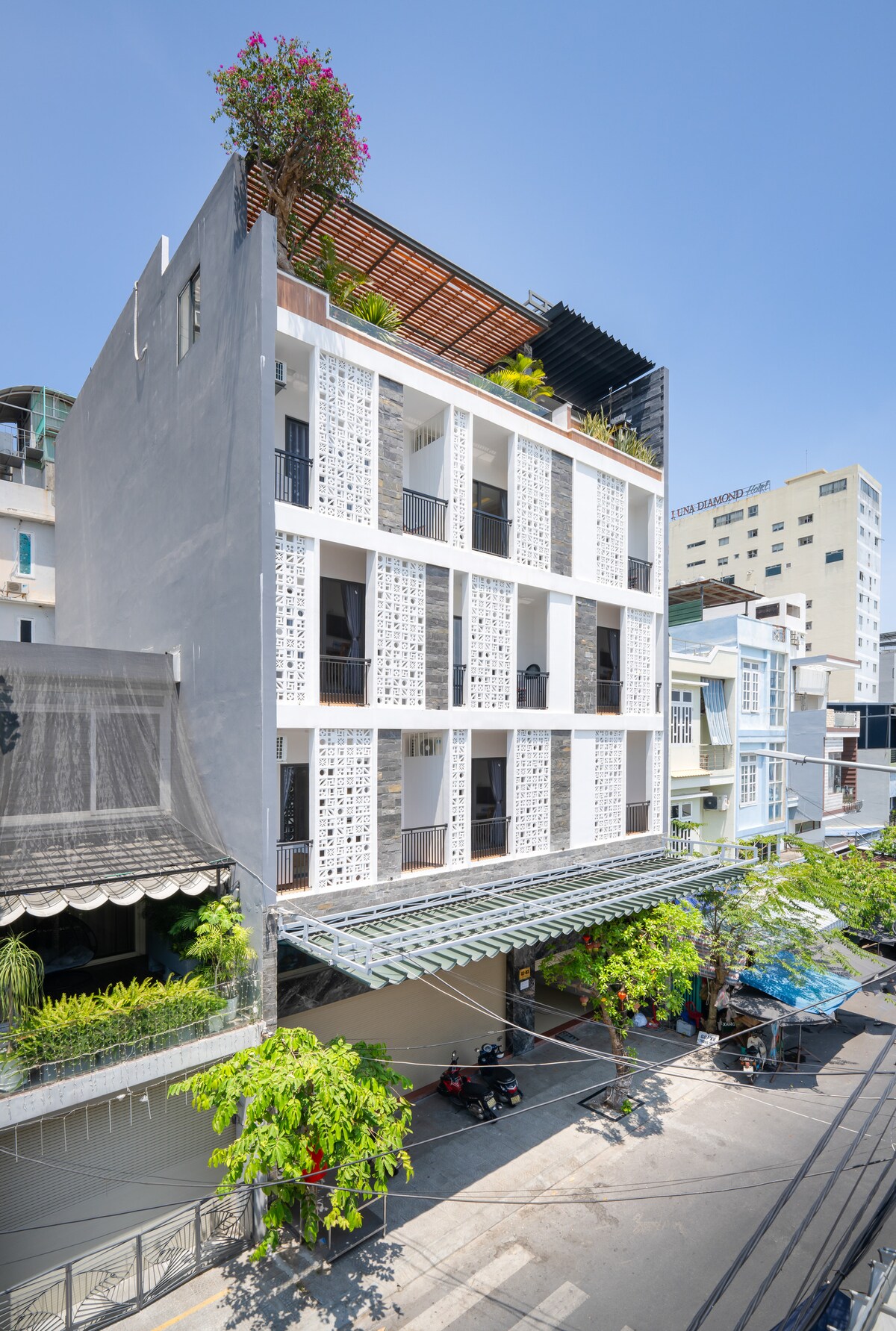 A modern multi-story building stands prominently, featuring a sleek facade with geometric wall patterns. Balconies adorned with greenery add a touch of nature. The surrounding area is lively, with trees lining the street and motorbikes parked nearby.