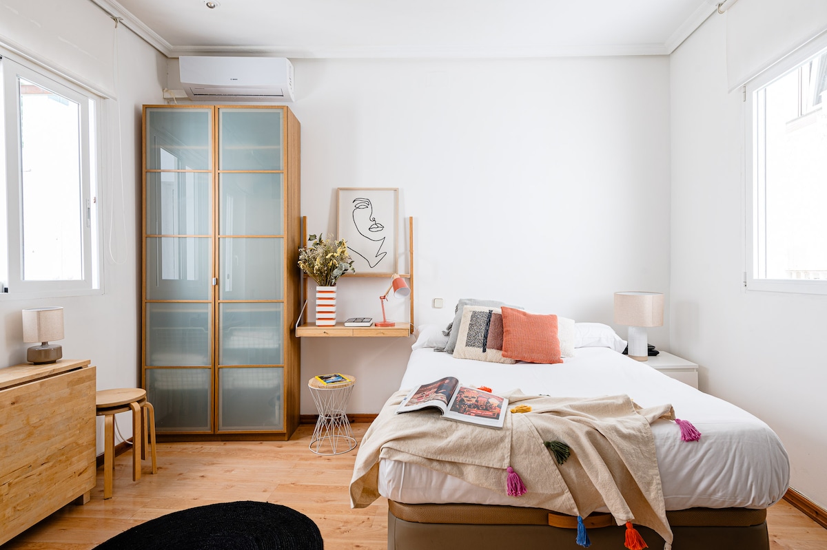 A bright room features a spacious bed with light bedding, complemented by colorful accent pillows. A minimalist wooden dresser and a small stool are present, alongside a cabinet with multiple glass doors. Natural light streams through two windows, enhancing the overall brightness of the space.
