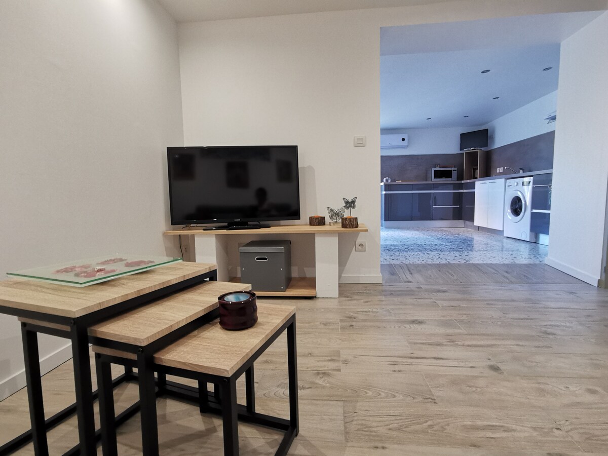 A cozy living area is presented with a television mounted on a wooden stand, accompanied by a set of nested tables. The space features light-colored wooden flooring and leads to a modern kitchen visible in the background, which is equipped with various appliances.