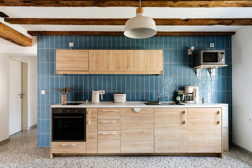 A modern kitchen features light wooden cabinetry complemented by a blue tiled backsplash. Counter space is organized with essential appliances, including an oven and microwave. Exposed wooden beams and a pendant light add character to the inviting space. Natural light brightens the room through an open doorway.