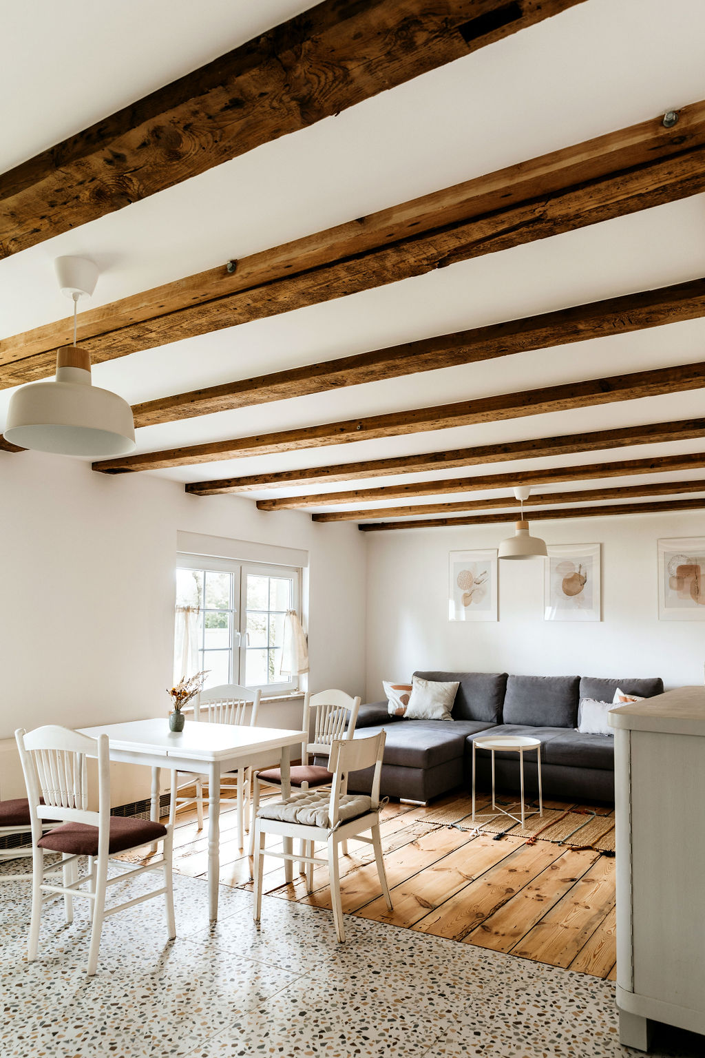 A spacious living area is highlighted by wooden beams and a mix of flooring. A gray sofa provides comfortable seating, accompanied by a white dining table with four chairs. Natural light filters through the window, enhancing the minimalist decor accented by framed artwork.