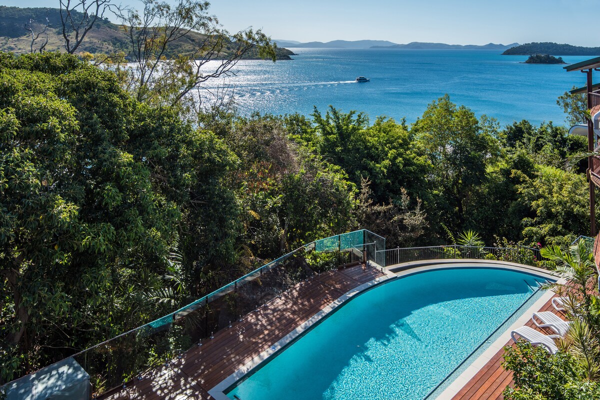 A clear view of a refreshing swimming pool surrounded by lush greenery, with the shimmering Coral Sea in the background. The expansive water glistens under the sunlight, and distant islands are visible on the horizon, enhancing the scenic landscape.
