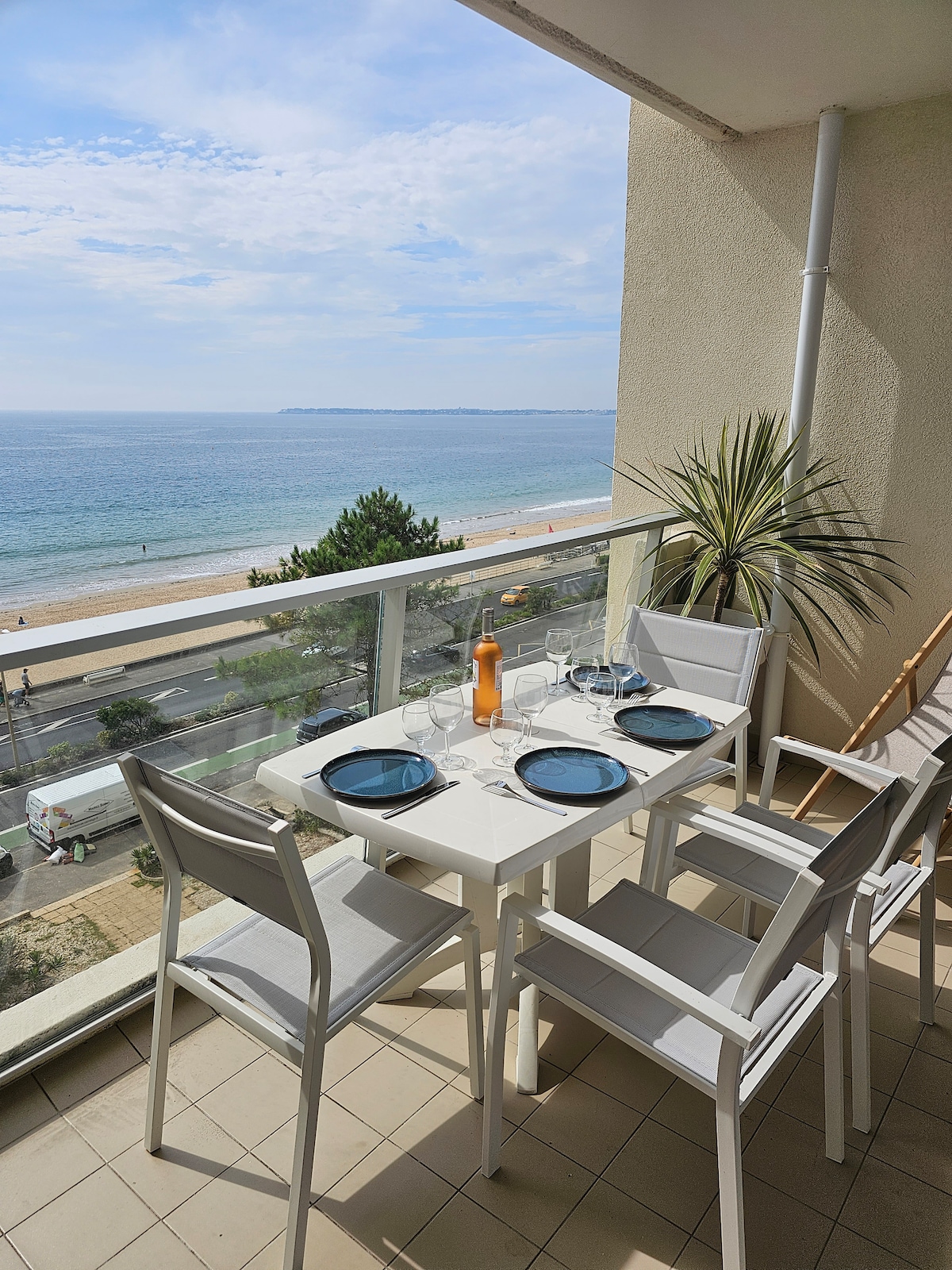 A balcony dining area features a white table set with blue plates and wine glasses, overlooking a scenic view of the ocean. Soft greenery is visible nearby, enhancing the outdoor space. The beach and promenade can be seen in the distance.
