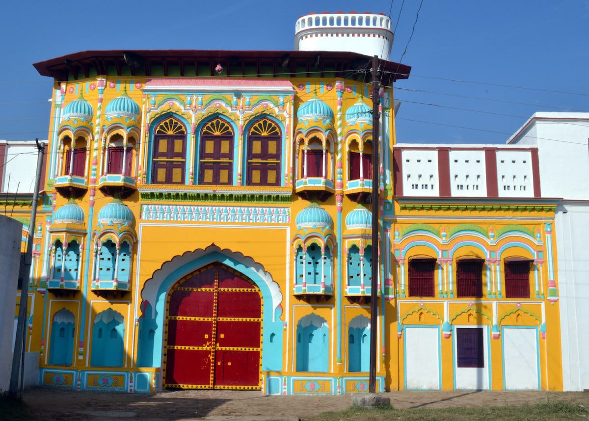 The vibrant façade of Haveli Jalpur features intricate designs, colorful arches, and decorative elements. A large red entrance door is framed by decorative patterns, while balconies adorned with blue domes offer an inviting glimpse of the historic exterior.