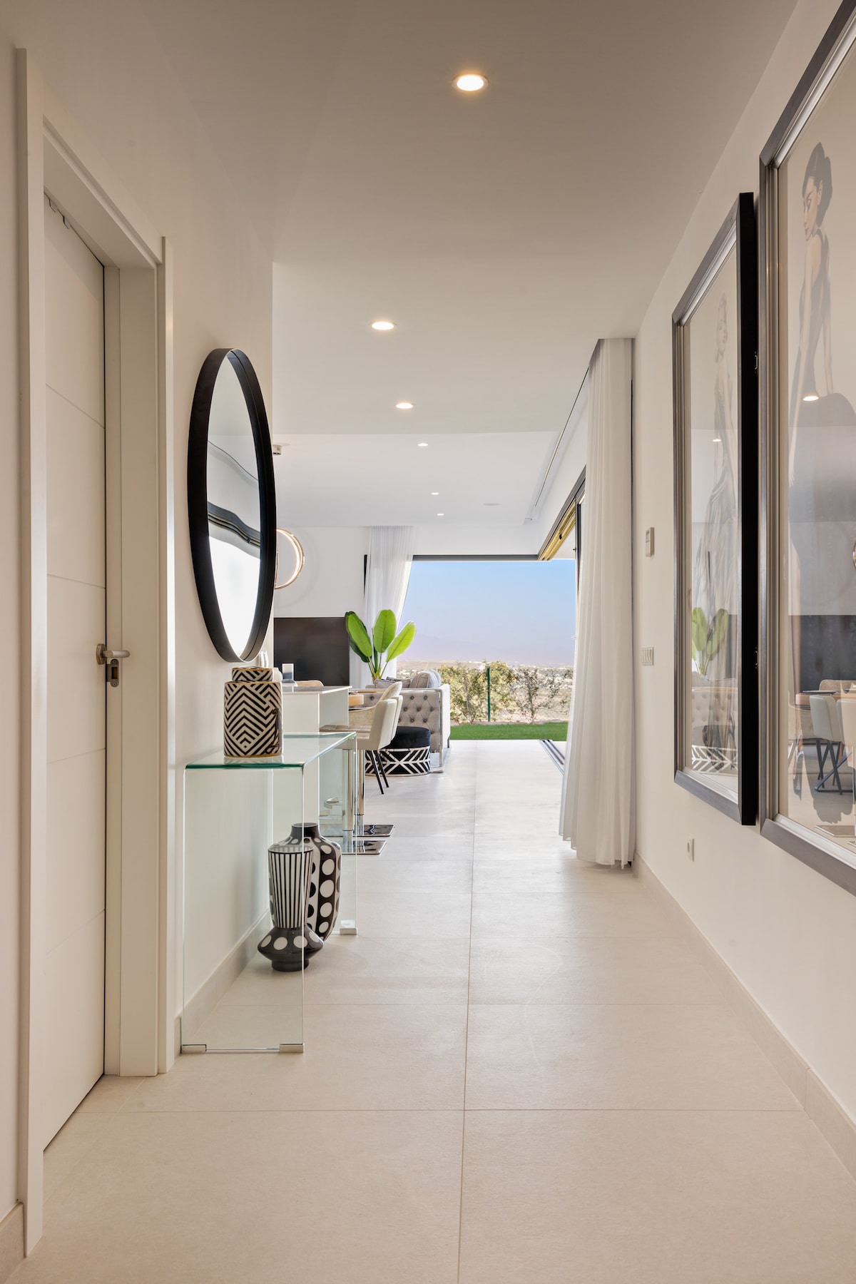 A bright hallway is seen featuring modern decor, including large framed artworks on the walls. A round mirror and a glass console table add to the contemporary design. Natural light flows in from an open area at the end of the hallway, highlighting the clean lines and tiled flooring.