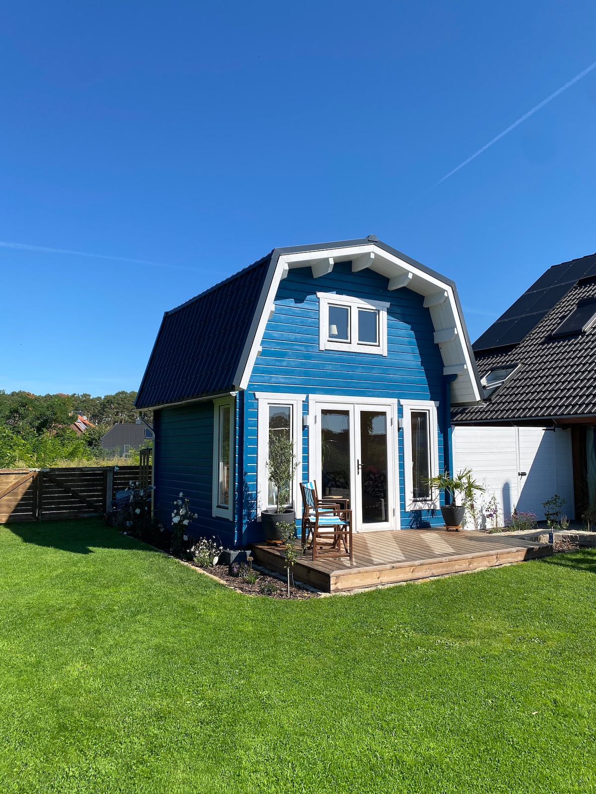 A charming blue tiny house is showcased against a clear blue sky, surrounded by a lush green lawn. A wooden deck extends from the entrance, adorned with potted plants and patio seating, inviting relaxation in a tranquil outdoor setting.