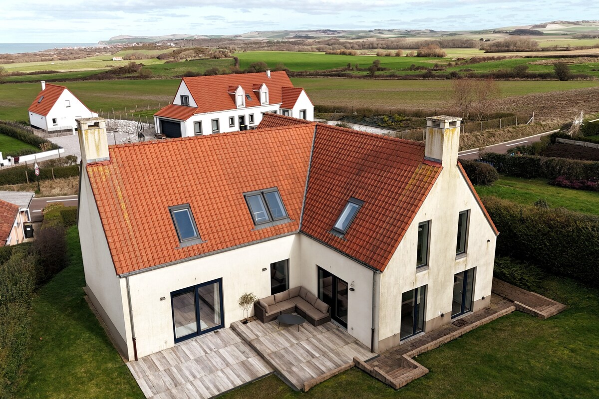 A modern house is presented from an aerial perspective, featuring a sloped roof with vibrant orange tiles. A spacious patio with outdoor seating is visible, surrounded by well-maintained greenery. The tranquil countryside and rolling hills extend in the background, enhancing the peaceful setting.