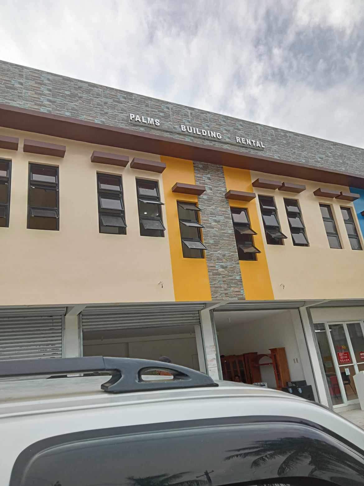 The facade of the Palms Building is displayed, featuring a mix of stone and painted surfaces. The building features several large windows that allow natural light to flow in, and a central sign indicating the name 'Palms Building Rental' above.