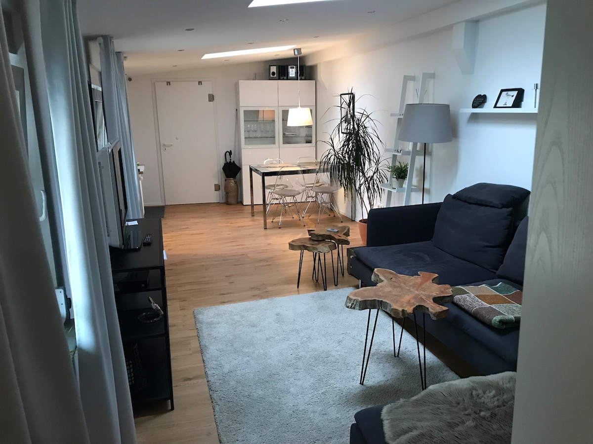 A bright living area features a dark sofa paired with unique wooden side tables. In the background, a dining area is visible, with a light-colored table and chairs beside a kitchen space. Natural light streams in through large windows, highlighting the warm wooden flooring.