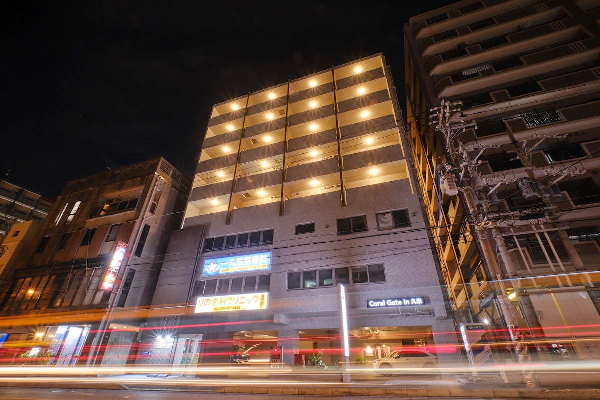 The exterior of a modern condominium is illuminated at night. The five-story building features a contemporary façade, with balcony railings visible on upper levels. Bright lights highlight the entrance, providing a welcoming appearance. The street is busy, with light trails suggesting movement nearby.