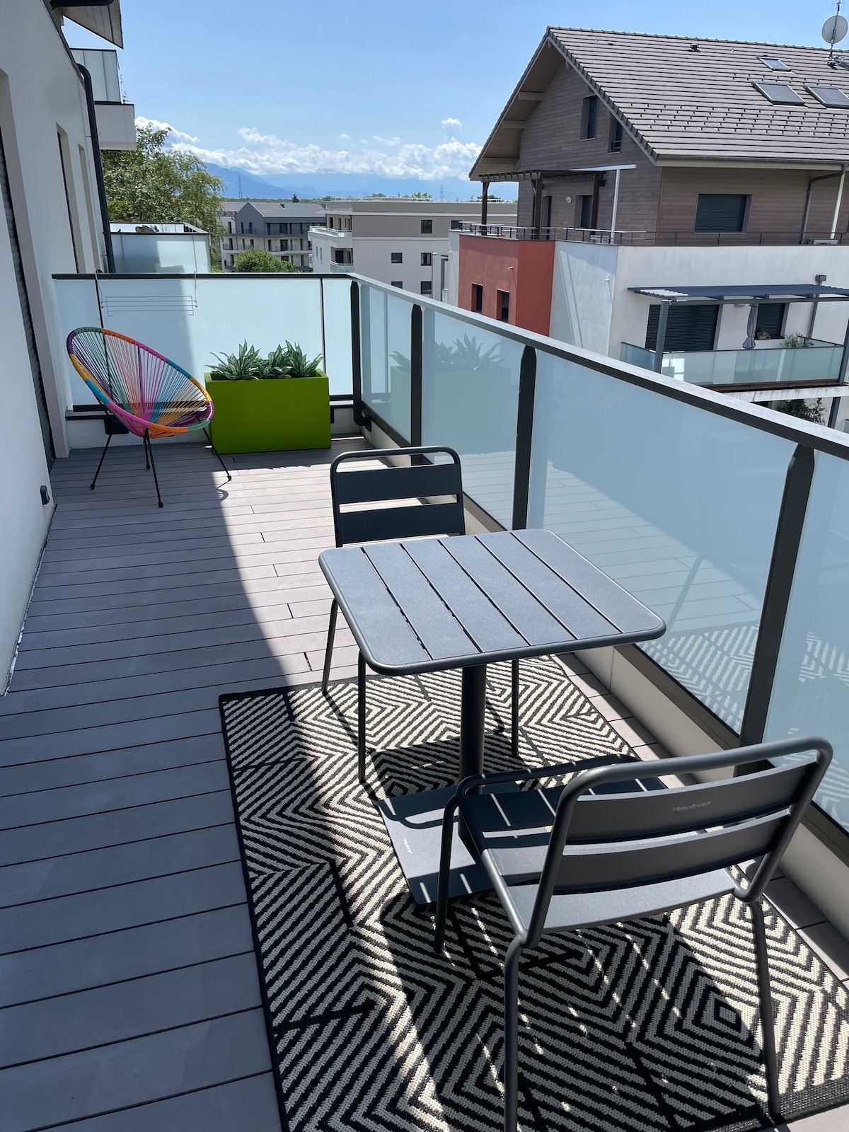 A private terrace is displayed, featuring a small table and two chairs positioned on a patterned rug. A colorful chair adds a touch of vibrancy. The glass railing provides a clear view of the surroundings, with distant mountains visible under a bright sky.
