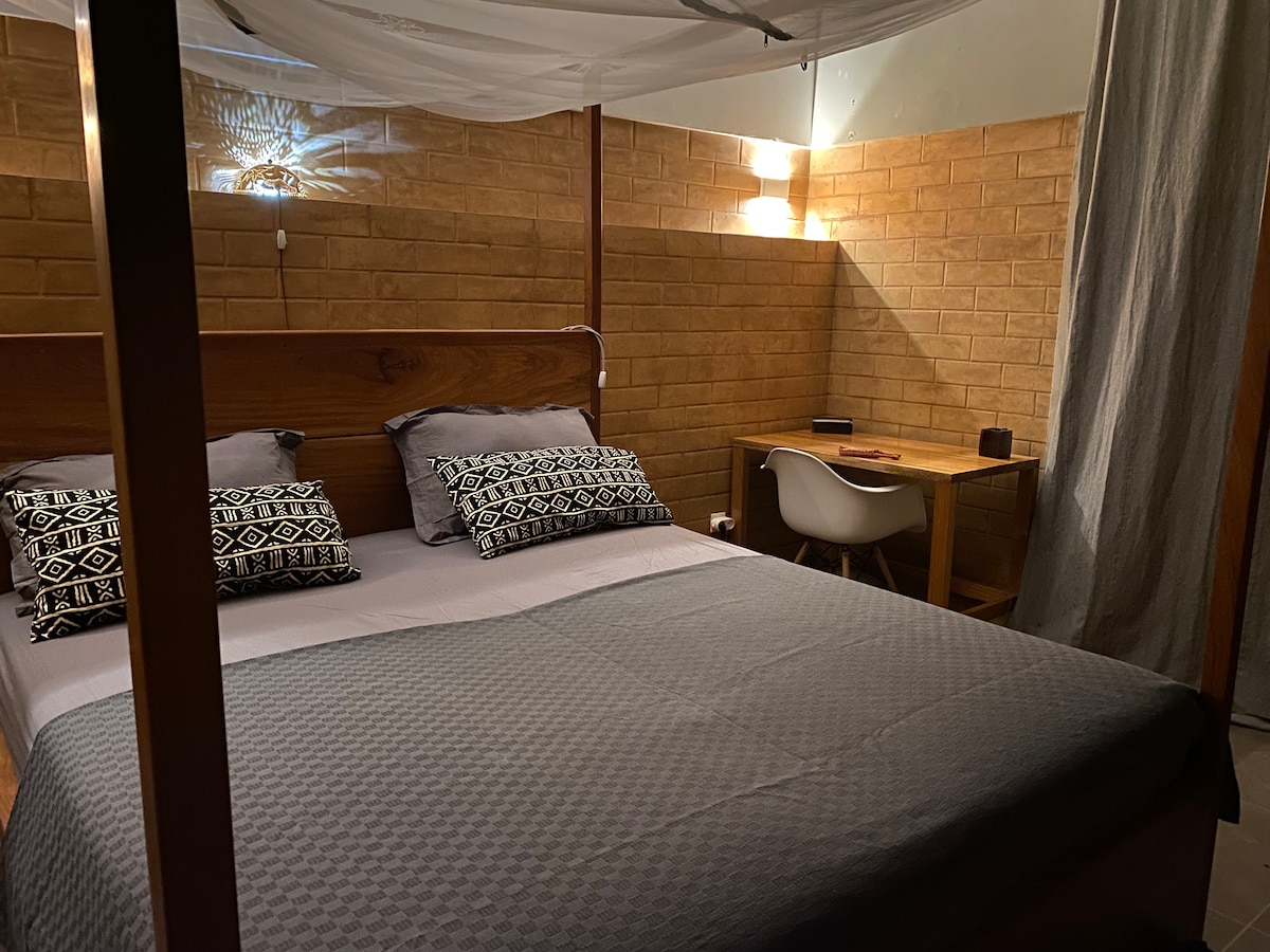 A comfortable bedroom features a spacious bed with a dark patterned duvet and several decorative pillows. A wooden desk and chair are positioned against a textured wall, providing a work area. Soft lighting enhances the room's calm atmosphere.