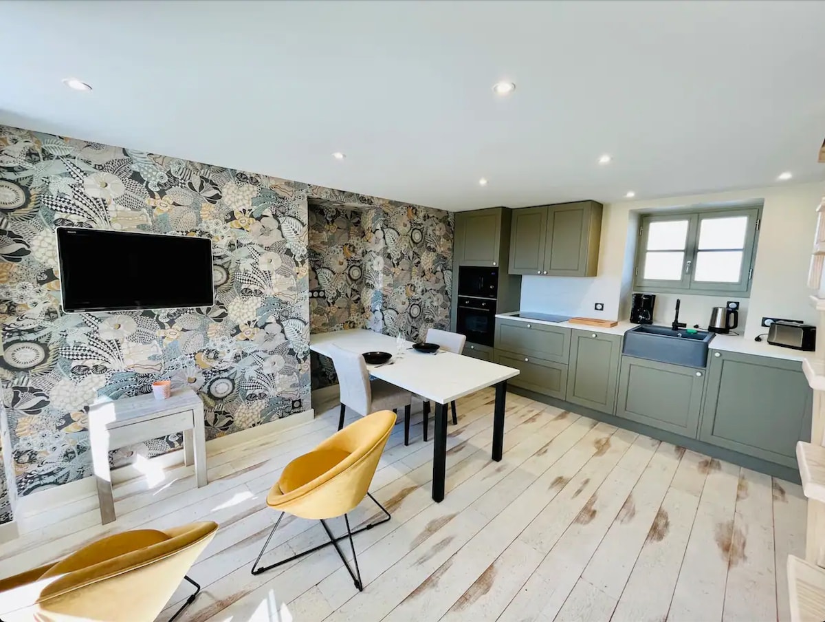 A bright living area features patterned wallpaper with floral designs, complemented by a white dining table and two stylish yellow chairs. A wall-mounted TV is visible, alongside modern kitchen amenities including an oven and coffee maker, all bathed in natural light from the nearby window.