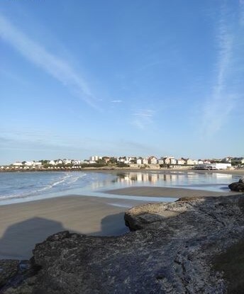 The image captures a serene beach scene, with gentle waves lapping at the sandy shore. In the background, a row of coastal houses is visible against a clear blue sky, reflecting on the water's surface. Rocky formations frame the foreground.