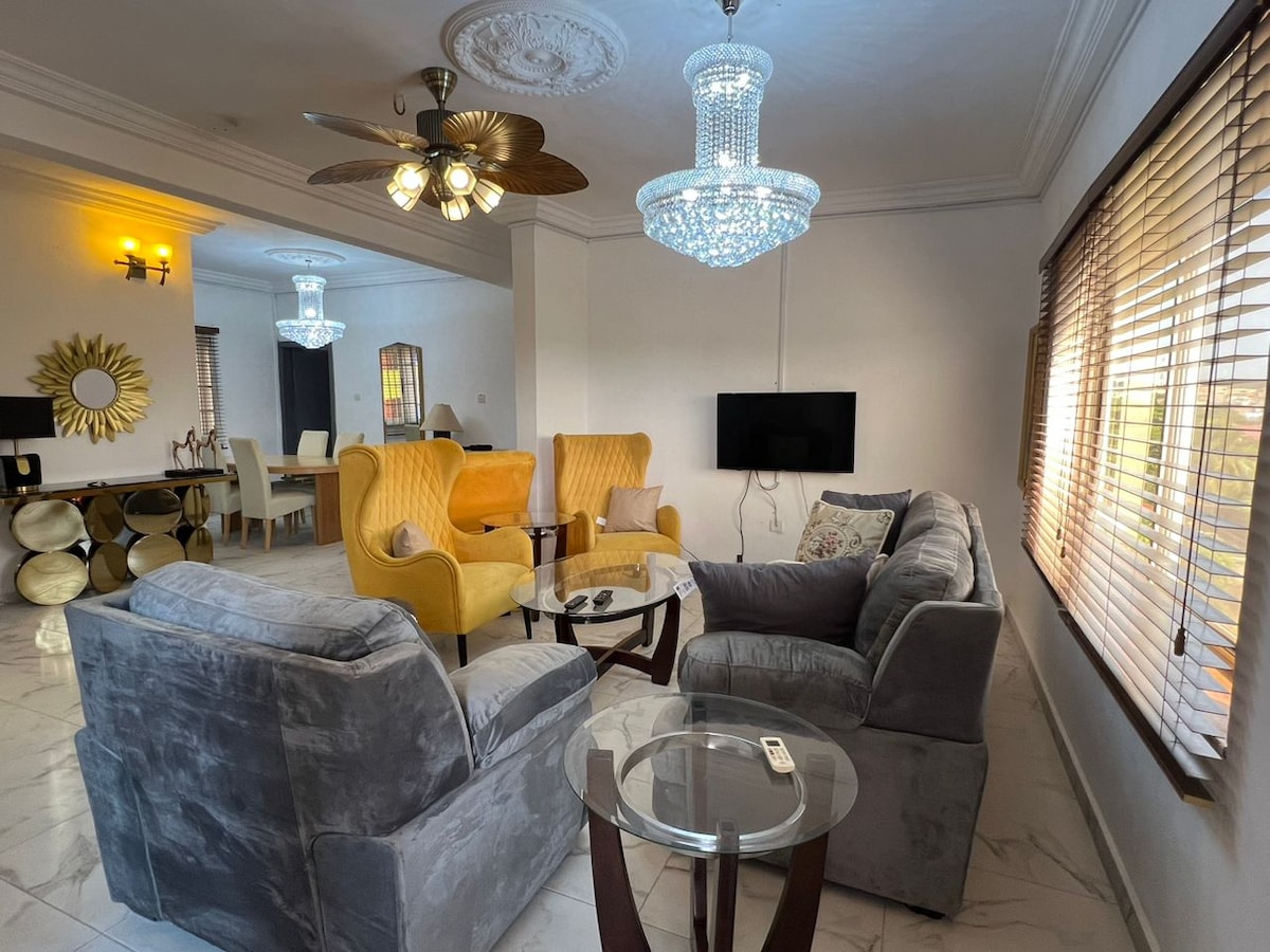 A modern living area is highlighted by elegant lighting fixtures and soft furnishings. Two yellow armchairs complement a plush gray sofa, arranged around a glass coffee table. Natural light filters through the window, and decorative mirrors reflect the space's stylish ambiance.