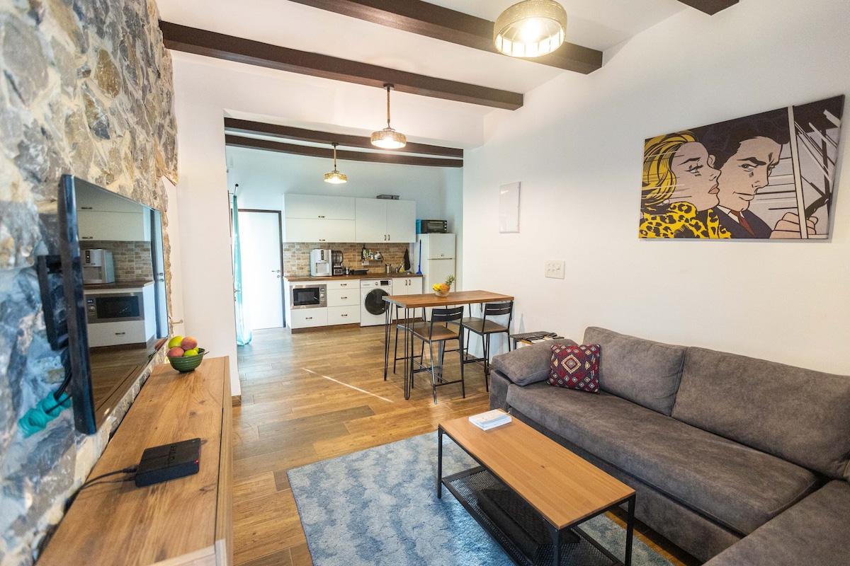 The interior showcases a cozy living area with a sectional sofa, a wooden coffee table, and a television mounted above a media console. A dining table for four is seen adjacent to an open kitchen, featuring wooden beams and a combination of stone and modern finishes. Natural light enters through a doorway leading outside.