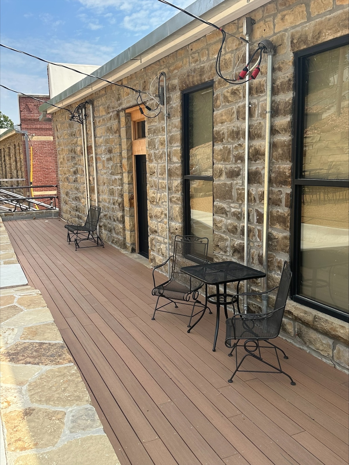 A private patio area is shown, featuring a wooden deck with two chairs and a small table. The stone wall of the building stands in the background, complemented by large windows. Natural light brightens the space, creating a relaxed outdoor setting.