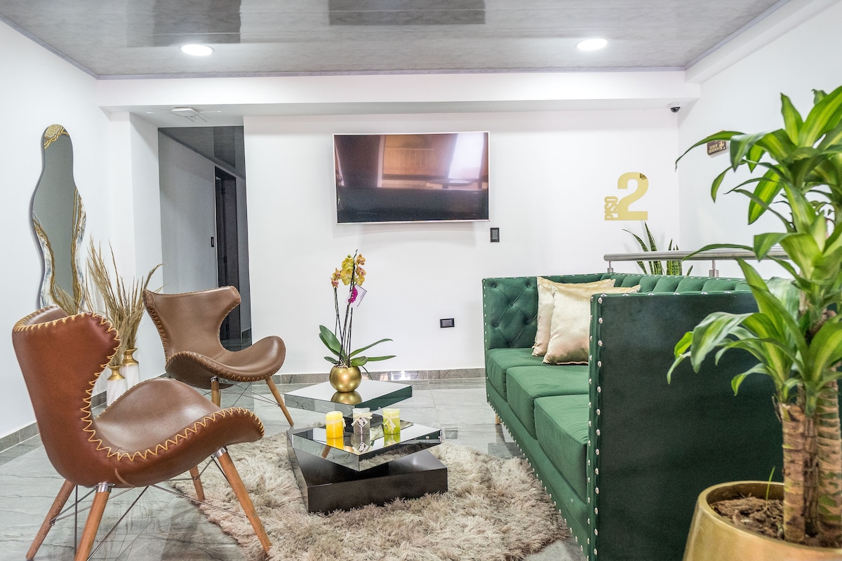 A modern living area features a green sofa, complemented by two brown accent chairs. A glass coffee table sits on a soft area rug, surrounded by decor including plants and flowers. A flat-screen TV is mounted on the wall opposite the seating arrangement.
