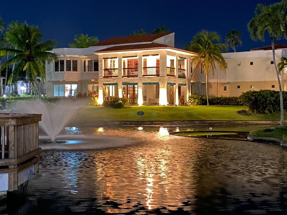 A well-lit two-story residence stands by a tranquil pond at night. Reflections of the building and lights shimmer on the water's surface, enhanced by a fountain in the foreground. Lush greenery and palm trees frame the scene, creating a serene tropical ambiance.