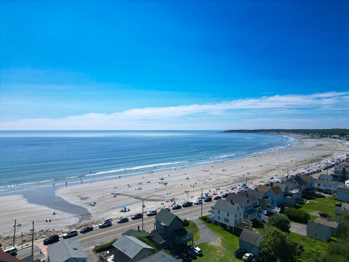 An aerial view captures the expansive Long Sands Beach stretching along the coast. Clear blue waters reflect the sky, while sunbathers and beachgoers enjoy the sandy shore. Nearby homes line the beach, offering a glimpse into this vibrant coastal community.