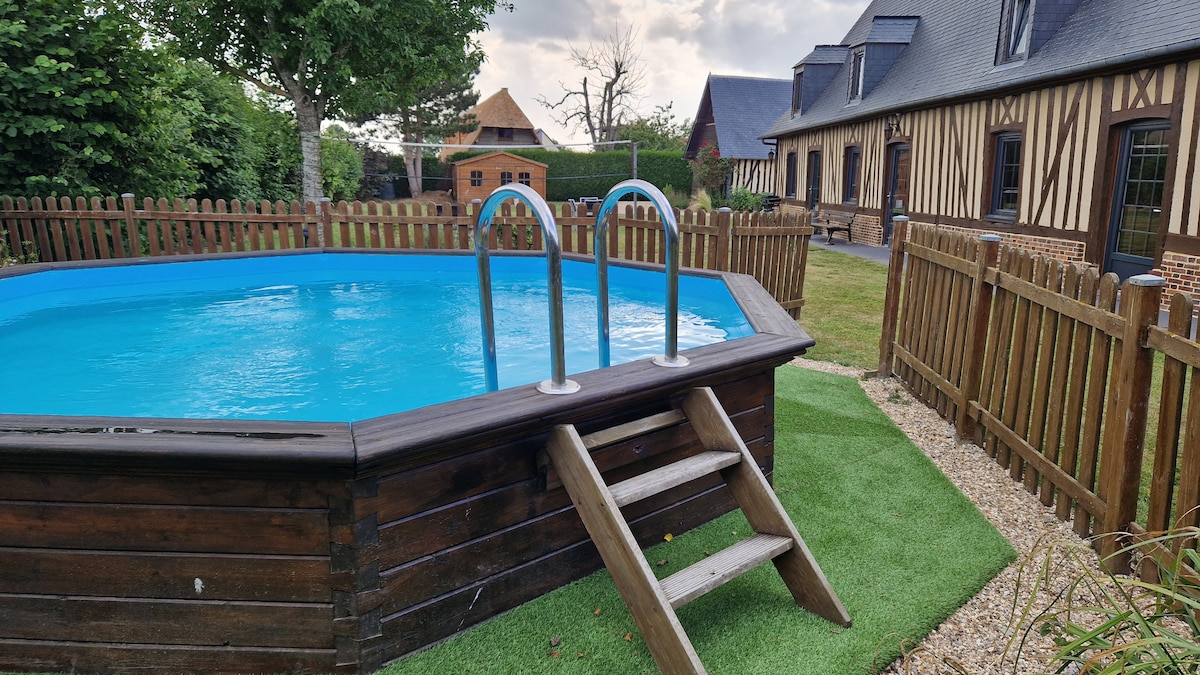 A semi-inground pool is featured in a well-maintained yard surrounded by a wooden fence. Ladders are positioned at one end for easy access. Lush greenery and manicured grass area complement the tranquil setting, with charming timber-framed buildings visible in the background.