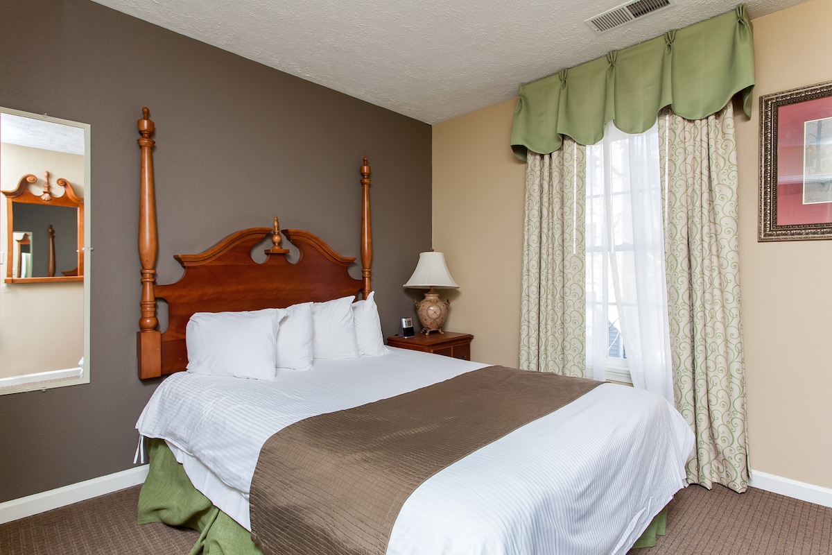 A well-furnished bedroom is showcased, featuring a spacious bed with white linens and a brown bedspread. Natural light enters through a window dressed in light green curtains. A mirror and a bedside lamp add functional elements, enhancing the room's overall appeal.