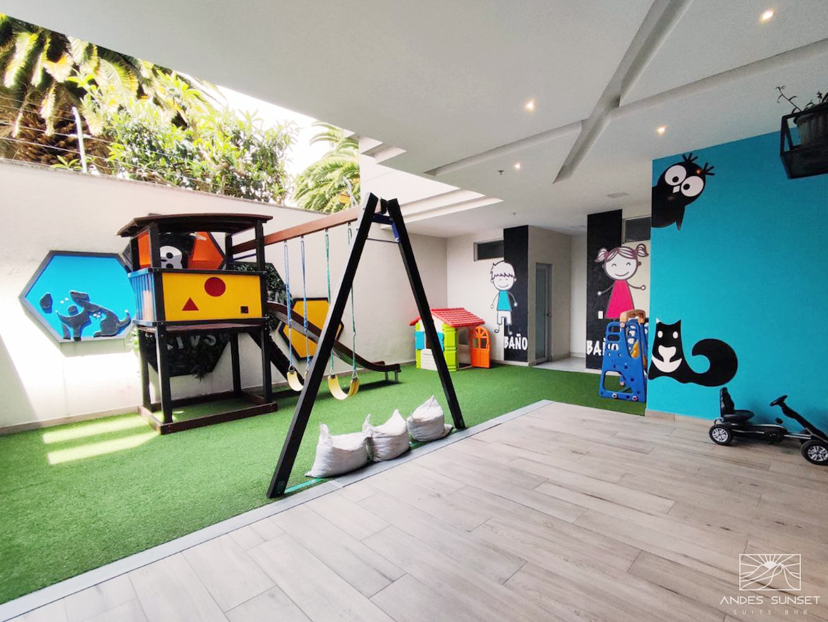 An engaging children's play area features a colorful play structure with slides and climbing elements, alongside black swings. Soft green grass creates a safe surface, complemented by playful wall graphics of animals and characters. Seating areas with bean bags and a small ride-on toy enhance the space.