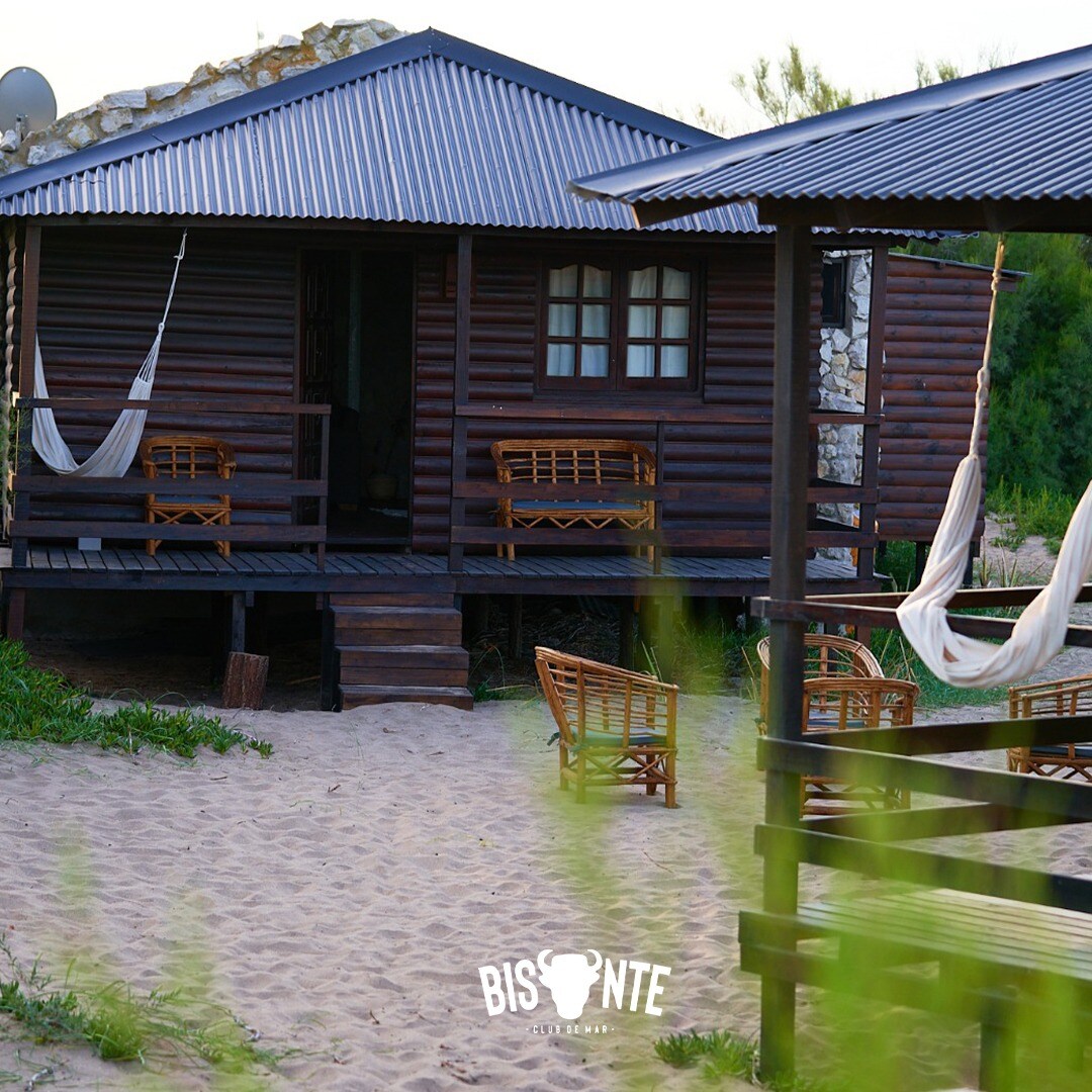 A wooden cabin features a porch with comfortable seating and hammocks. The structure blends with sandy surroundings, surrounded by lush greenery. Large windows provide natural light, and the rustic design enhances the connection with the natural environment.