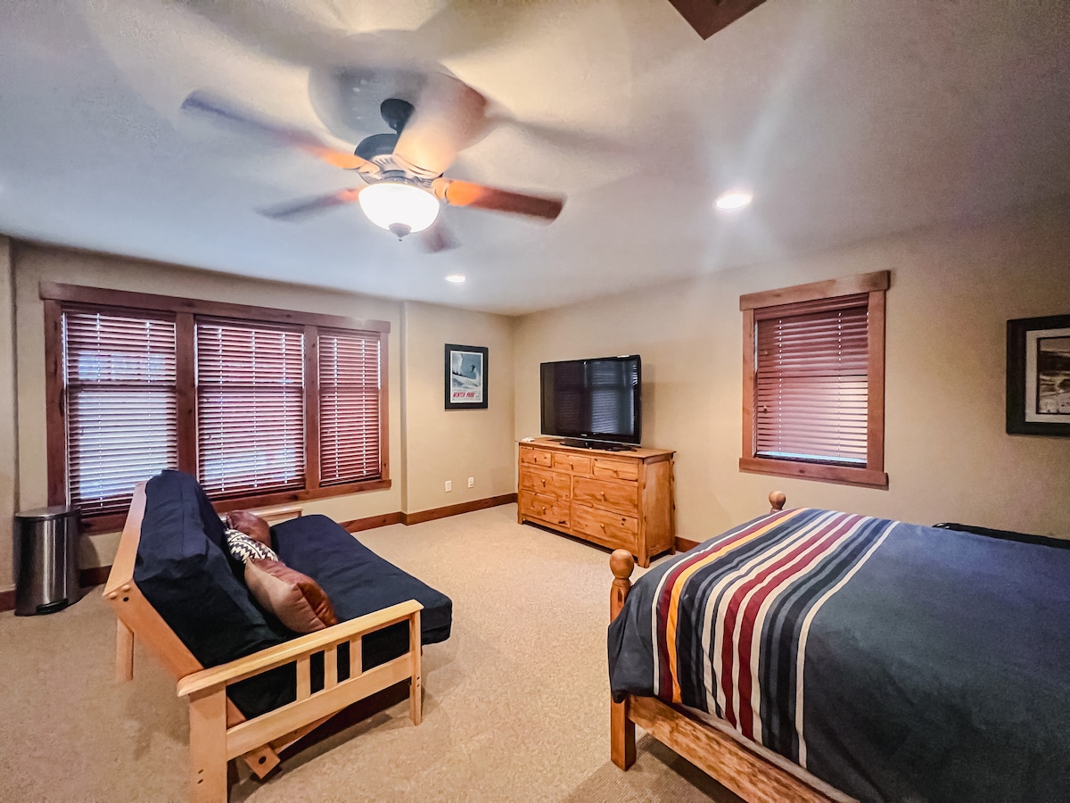 A spacious studio features a queen-sized bed with a striped blanket and a queen-sized futon against a wall. Natural light fills the room through two large windows with wooden blinds. A flat-screen TV is mounted above a wooden dresser, providing a cozy atmosphere.