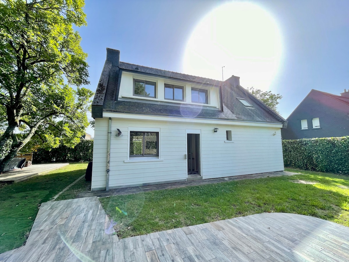 The exterior view of the house is presented, showcasing its white wooden siding and sloped roof. A spacious lawn is visible, bordered by lush greenery, with a patio area partially shown under bright sunlight.