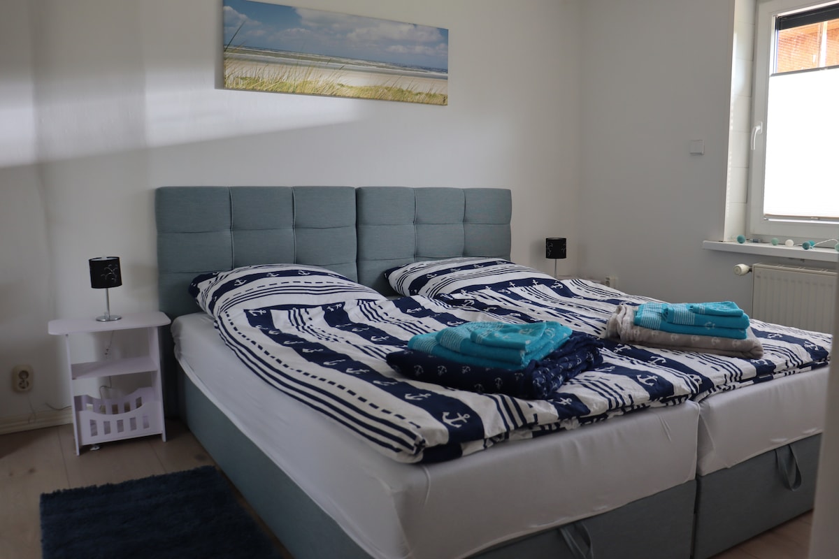 A serene bedroom features a spacious bed with separate mattresses, dressed in nautical-themed bedding. Two bedside lamps provide soft lighting, while a window allows natural light to filter into the room. Fresh towels are neatly arranged for convenience.