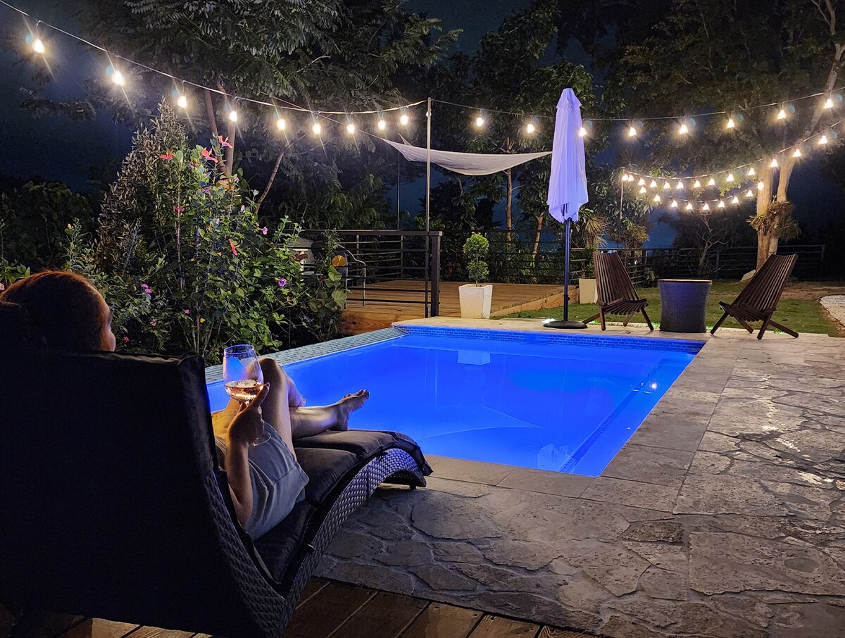A serene outdoor area is illuminated by string lights, surrounding a private pool that glows in soft blue. Comfortable loungers provide seating, and lush greenery enhances the ambiance. The scene captures a peaceful evening setting, perfect for relaxation.