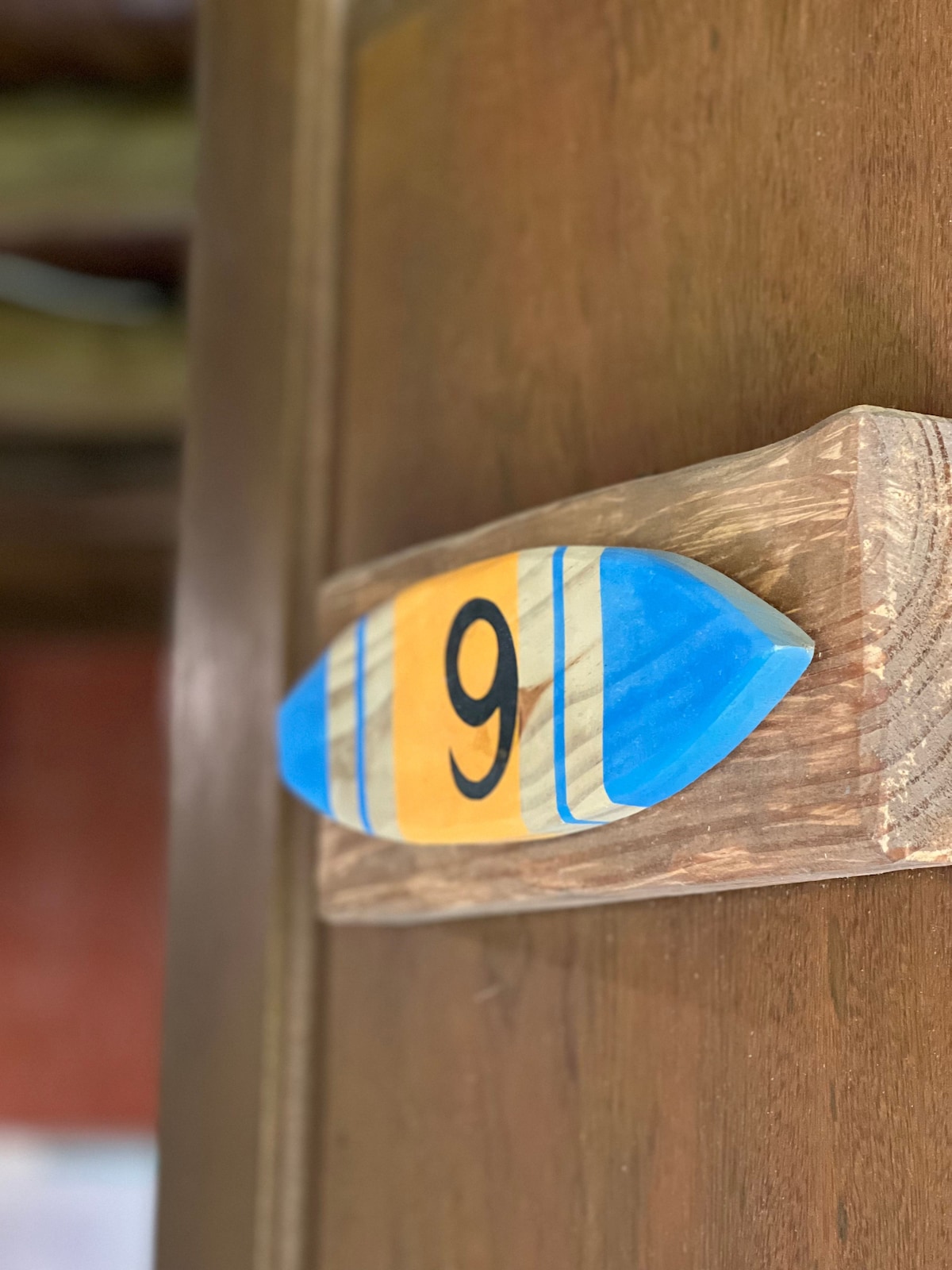 A wooden door sign is displayed, featuring a blue and yellow surfboard design with the number nine prominently painted in black. The texture of the wood is visible, suggesting a rustic charm.