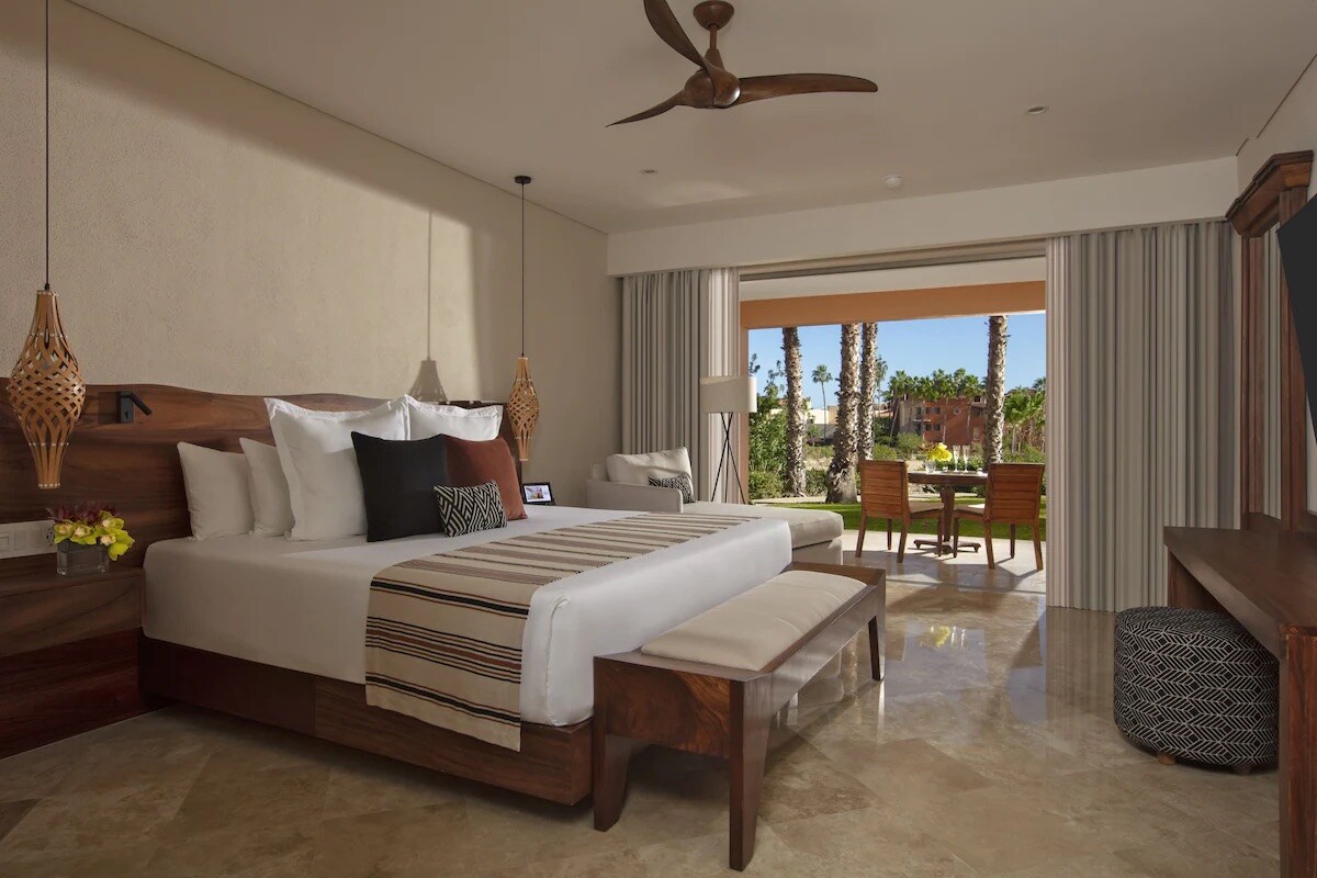 A spacious bedroom features a large bed with multiple pillows, complemented by a decorative bench at its foot. Natural light enters from the large sliding door, leading to an outdoor seating area with views of palm trees and greenery. Elegant light fixtures add a modern touch.