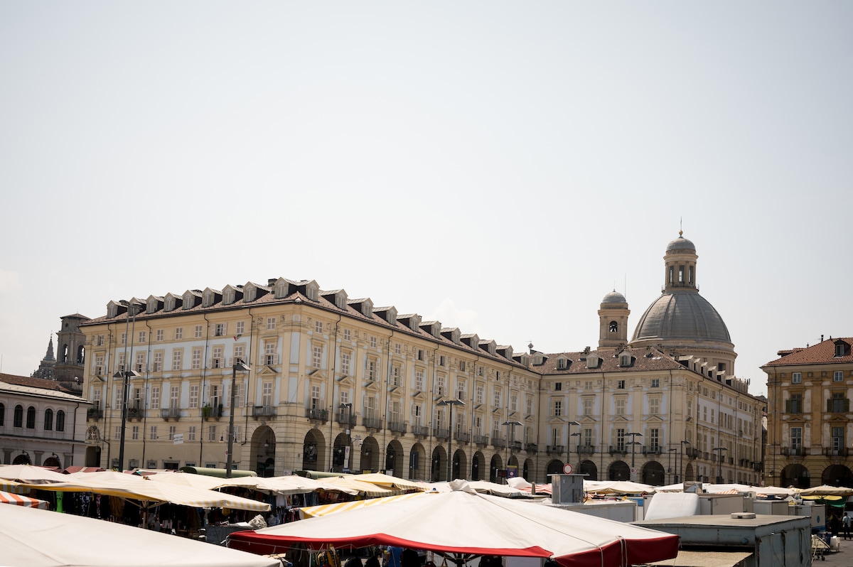 A historic building with a light-colored facade and arched columns is prominently featured. The structure includes large windows and a notable dome atop the roof. Market stalls are set up in the foreground, offering a variety of goods against the backdrop of the vibrant square.