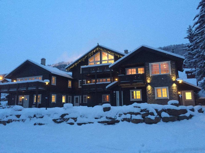 An elegant mountain lodge is surrounded by snow, with warm light emanating from its windows. The structure features a combination of wooden and stone elements, while festive lights adorn the roofline. Snow blankets the ground and nearby surfaces, providing a serene winter setting.