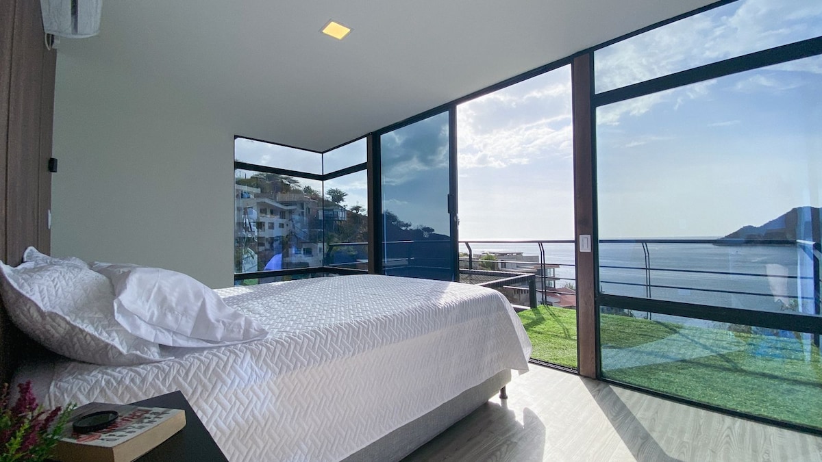 A bright bedroom features a spacious bed covered with a light quilt, positioned near large glass windows. These windows offer panoramic views of the sea and coastal landscape. Natural light fills the space, and a small bedside table is visible in the foreground.