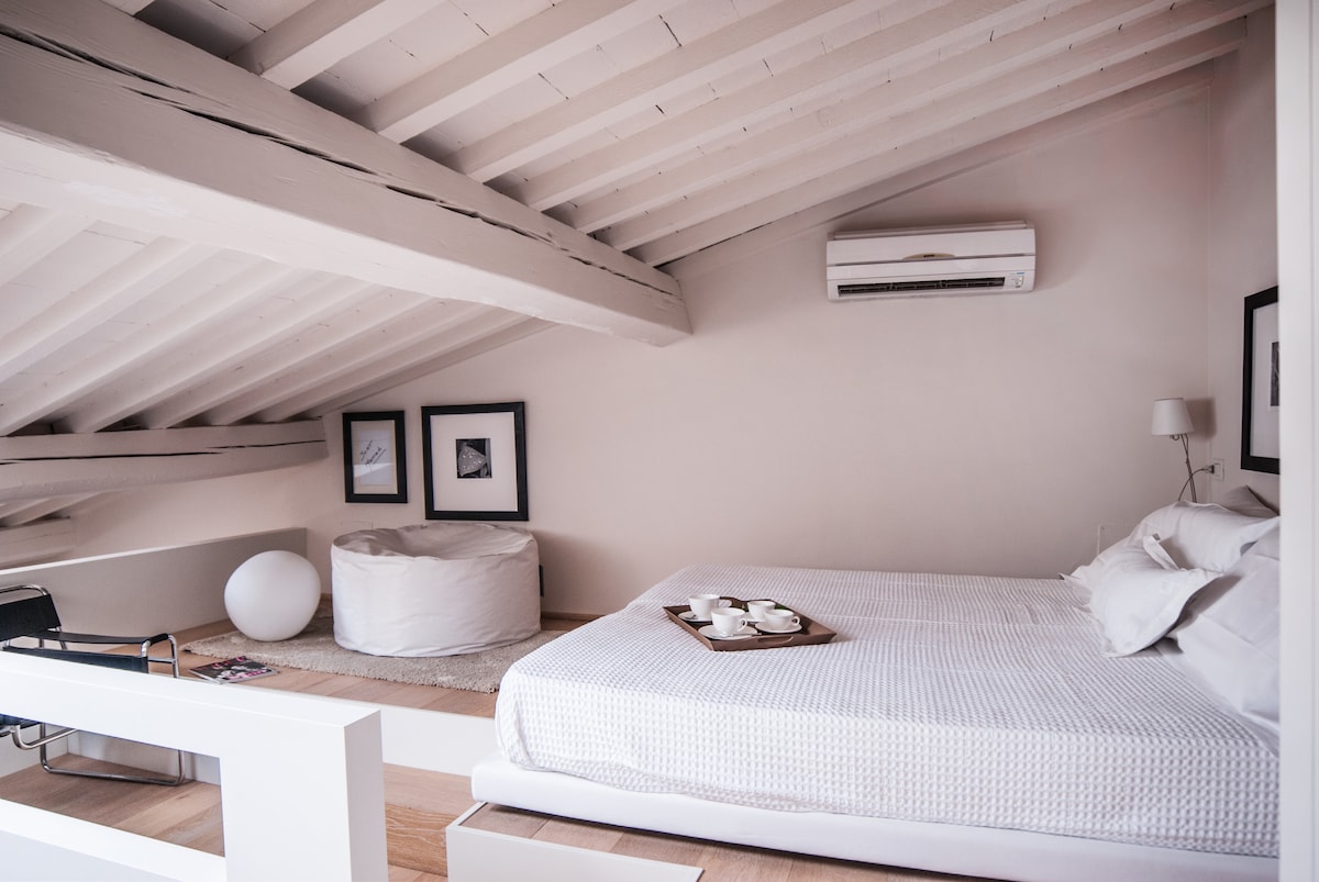 The spacious loft area features a large bed with a white textured coverlet, flanked by minimalist art on the walls. An air conditioning unit is positioned above, while a circular ottoman and a small table are visible. Natural light filters softly through the room.