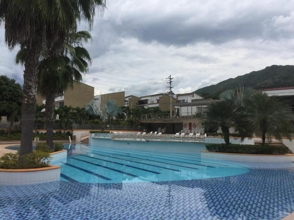 A serene swimming pool is surrounded by palm trees and well-kept landscaping. The pool features blue-tiled edges, with gentle ripples reflecting the cloudy sky. Chairs are arranged neatly along the perimeter, providing spaces for relaxation. Nearby, residential buildings and mountains create a scenic backdrop.