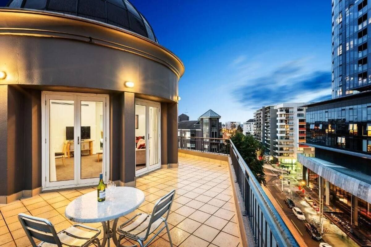 A spacious balcony is shown, featuring a round table with two chairs, overlooking the city lights. Large glass doors lead into the interior living area, while the skyline includes a mix of modern buildings and street activity, all under a twilight sky.