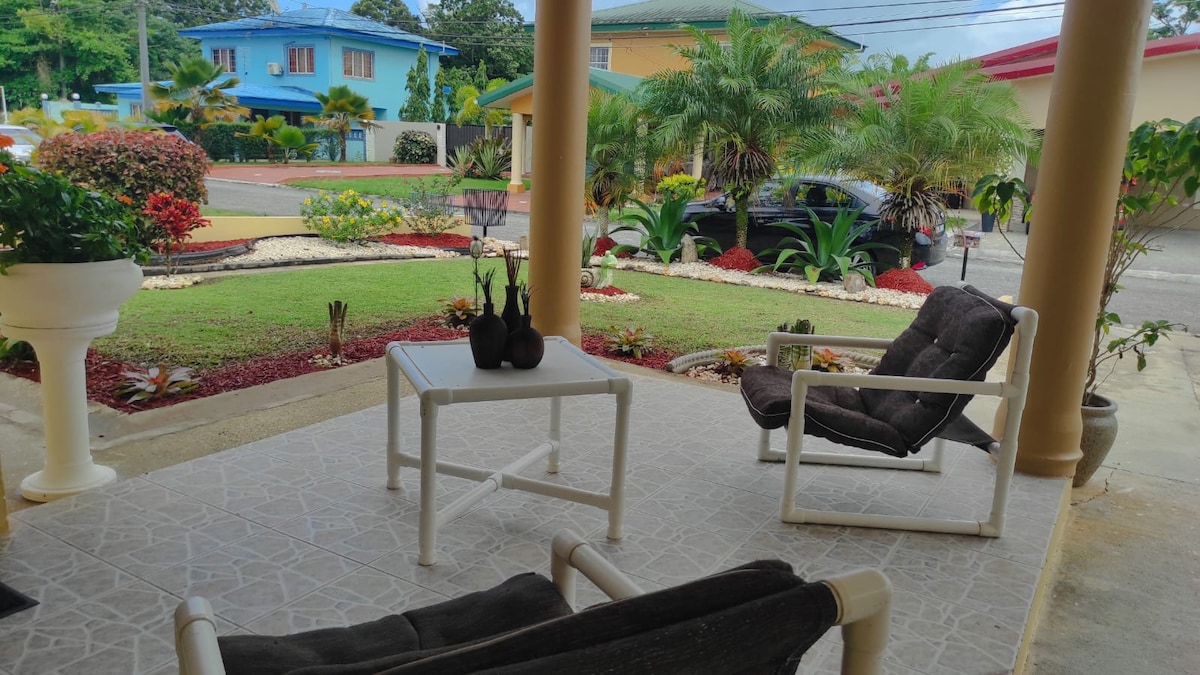 A covered outdoor area features two comfortable chairs and a small table. Lush greenery and colorful flowers surround the space, enhancing the welcoming entrance to the home. A parked vehicle is visible in the background, along with well-maintained landscaping.