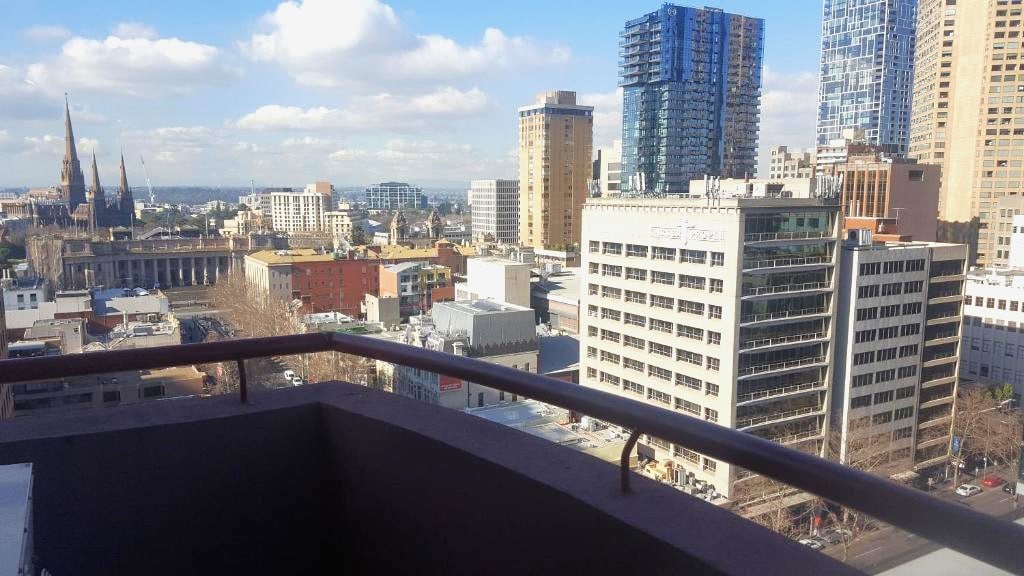 A spacious balcony offers views of the Melbourne skyline, featuring a mix of historic and modern buildings. Notable structures, including the spires of a nearby cathedral, are visible against a bright blue sky adorned with fluffy clouds.