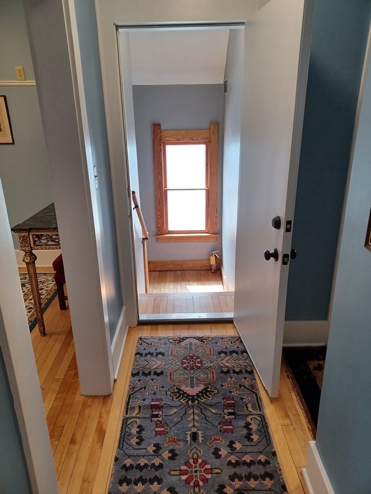 A well-lit entrance is framed by a closed door, revealing a hallway featuring warm wooden flooring and a decorative area rug. A window at the end of the hall allows natural light to brighten the space, while stairs lead to the upper level.