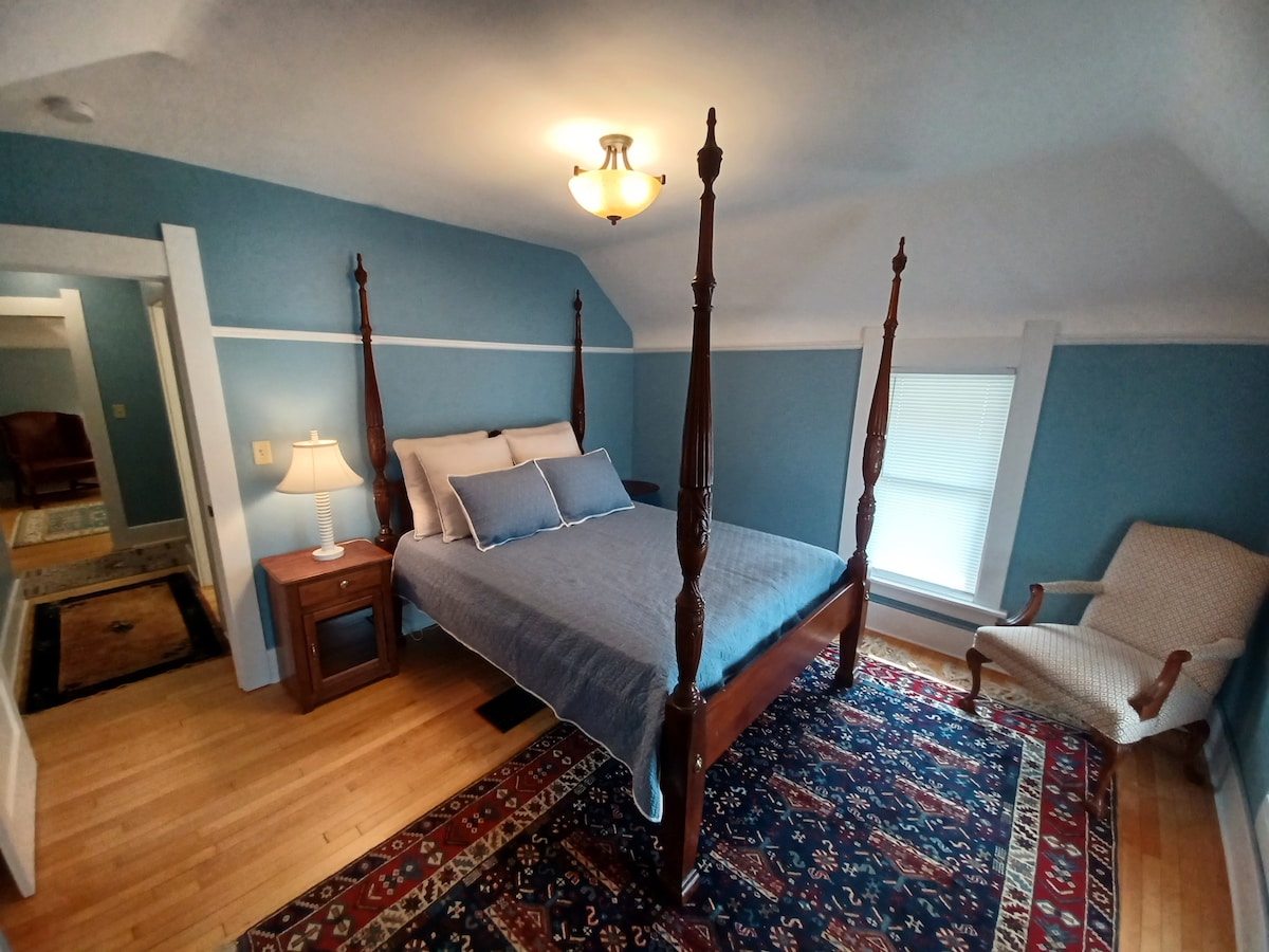 The room features a four-poster bed with a soft blue bedspread and accent pillows, complemented by a wooden nightstand with a lamp. A cozy armchair and a patterned area rug enhance the warm atmosphere, while natural light filters through the nearby window.