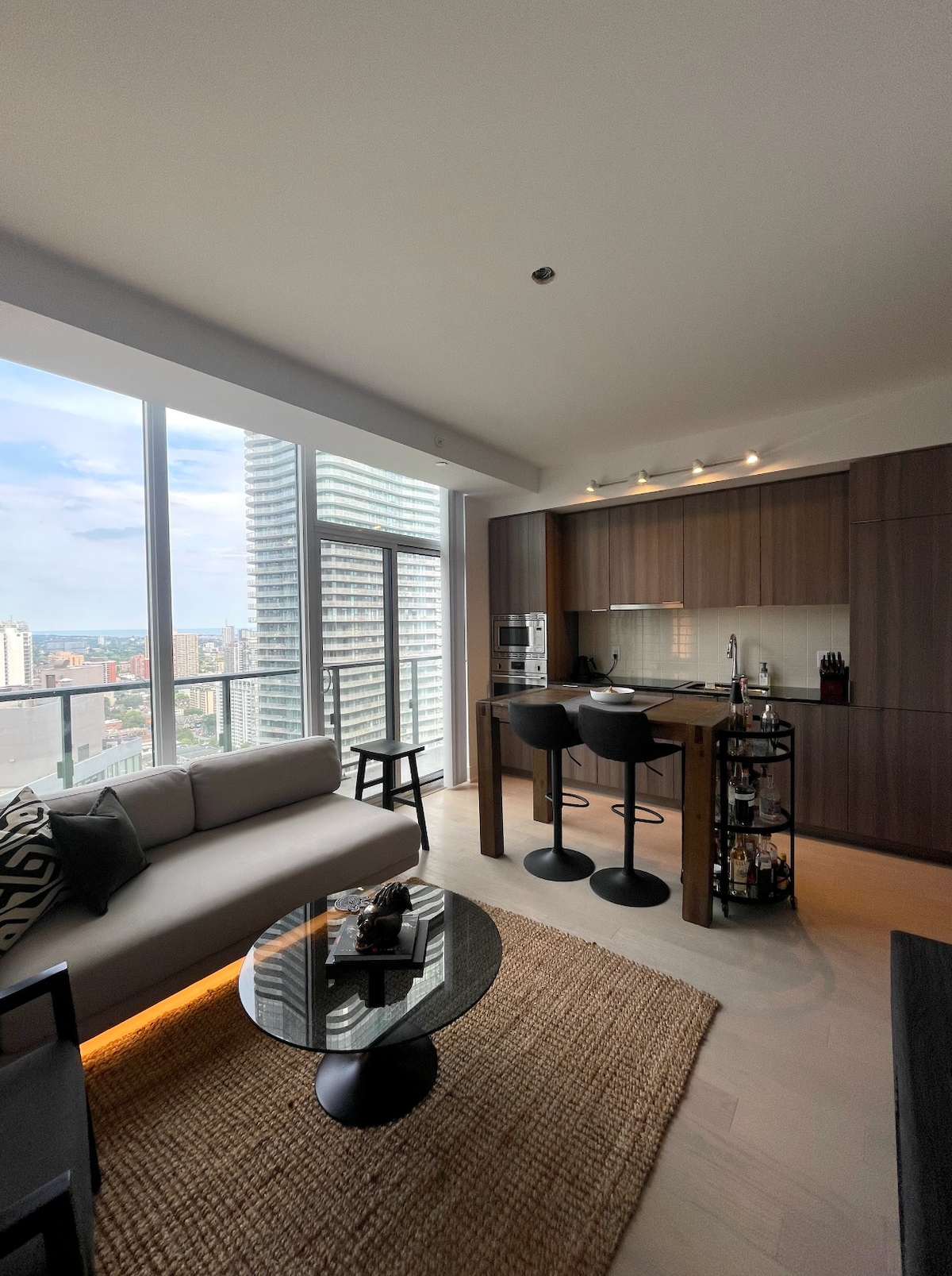 A modern living space features large windows that provide natural light and city views. A sleek kitchen with dark cabinetry is complemented by a round dining table with two stools. A cozy sofa sits beside a circular coffee table, all placed on a textured area rug.