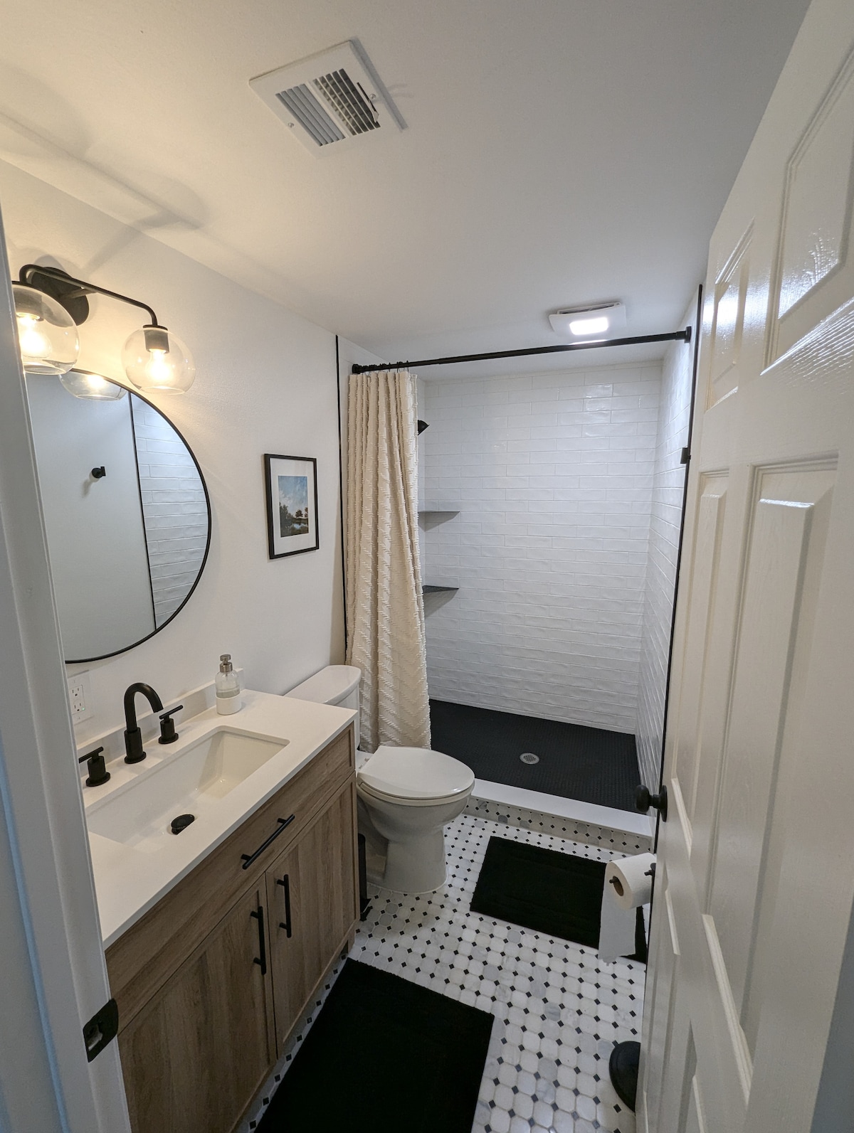 A well-designed bathroom features a modern sink with a spacious countertop and under-sink storage. A circular mirror reflects light above the sink. The shower area is enclosed with a white tile backdrop and includes shelves for toiletries. Contrasting black and white tiles cover the floor.